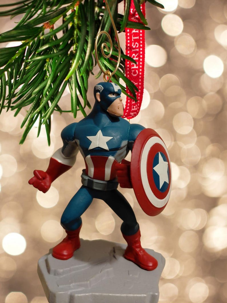 May include: A Captain America Christmas ornament. The figurine is in a heroic pose, holding his shield. The ornament is blue, red, and white, with a star on the shield and chest. It hangs from a gold wire and a red ribbon that says "MERRY CHRISTMAS".