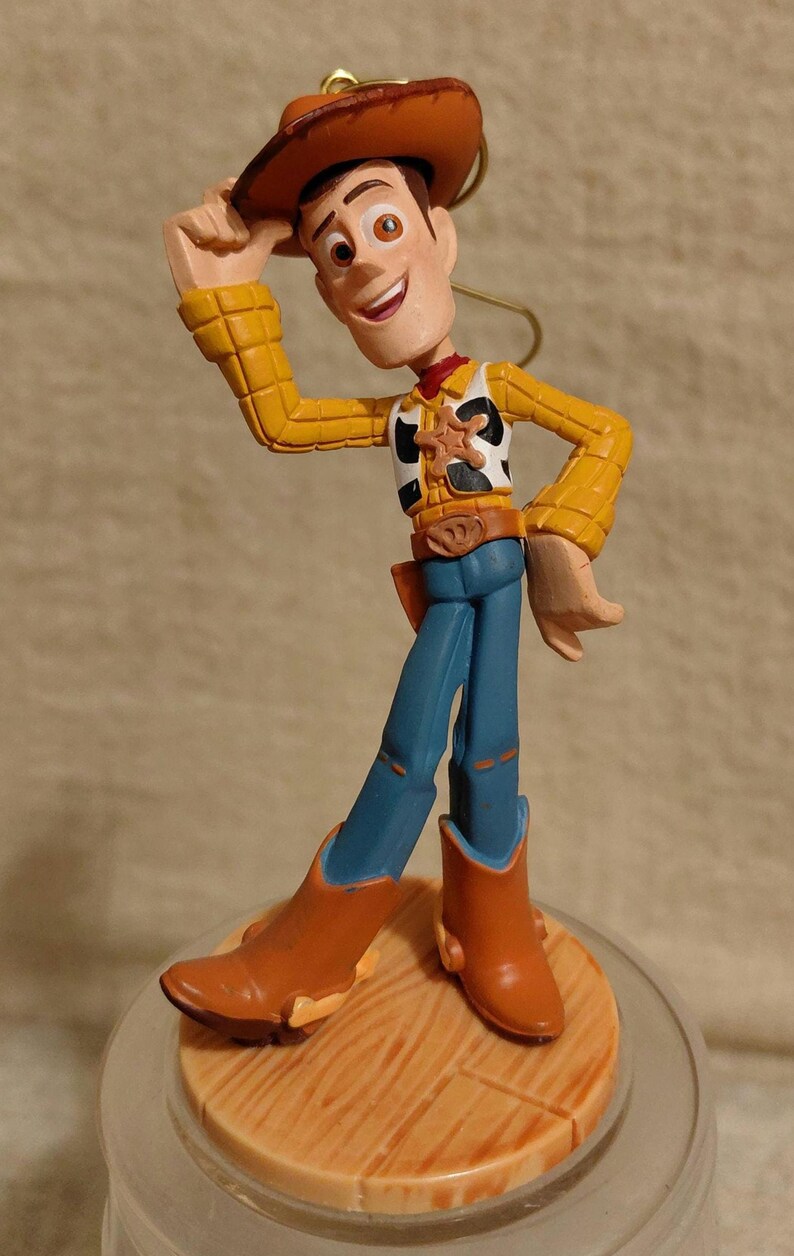 May include: A figurine of Woody from Toy Story, wearing a brown cowboy hat, yellow shirt, blue jeans, and brown boots. The figure is standing on a wooden-style base. The character is smiling and tipping his hat.