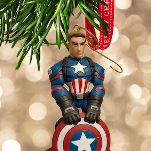 Puede incluir: Un adorno navide&ntilde;o de Capit&aacute;n Am&eacute;rica. La figura est&aacute; en una pose heroica, sosteniendo su escudo. El adorno es azul, rojo y blanco, con un colgador dorado y una cinta roja que dice "Merry Christmas".