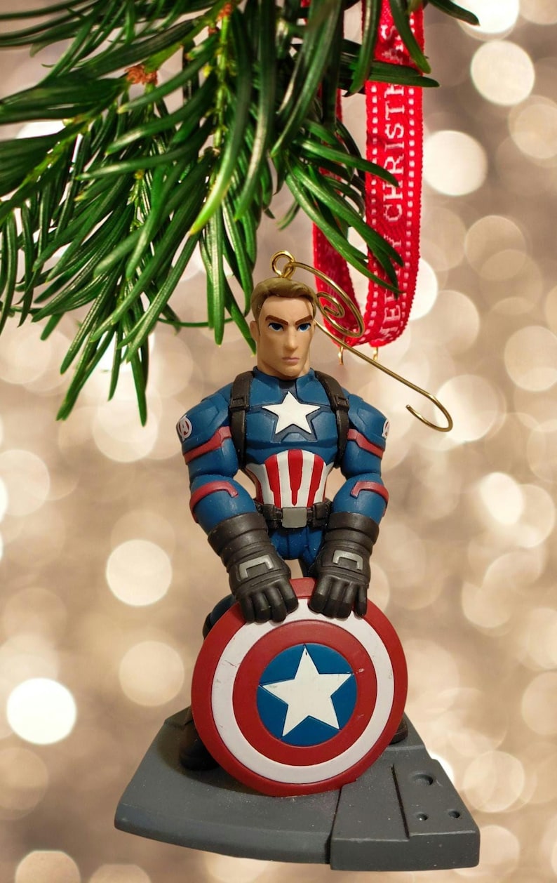 Puede incluir: Un adorno navide&ntilde;o de Capit&aacute;n Am&eacute;rica. La figura est&aacute; en una pose heroica, sosteniendo su escudo. El adorno es azul, rojo y blanco, con un colgador dorado y una cinta roja que dice "Merry Christmas".