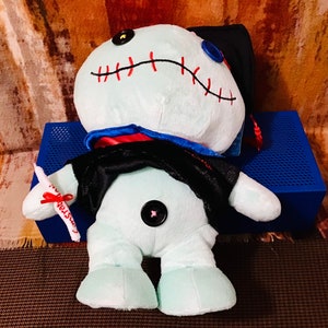May include: A light blue plush toy wearing a black graduation gown and cap. The toy has red stitching around the mouth and a black button nose. The toy is holding a white scroll with a red ribbon.