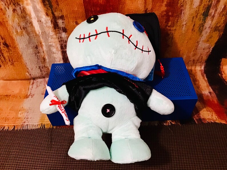 May include: A light blue plush toy wearing a black graduation gown and cap. The toy has red stitching around the mouth and a black button nose. The toy is holding a white scroll with a red ribbon.