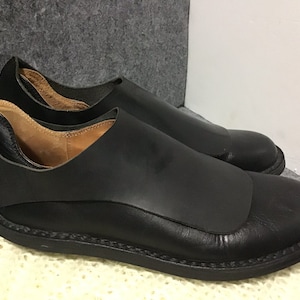 May include: A pair of black leather slip-on shoes with a unique design featuring a flap over the top of the foot. The shoes have a stitched sole and a rounded toe.