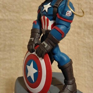 Puede incluir: Figura de Capit&aacute;n Am&eacute;rica, con un traje azul y rojo y una estrella blanca. La figura sostiene un escudo con una estrella blanca sobre un c&iacute;rculo azul, rodeado de anillos rojos y blancos. Con un lazo dorado para colgar.