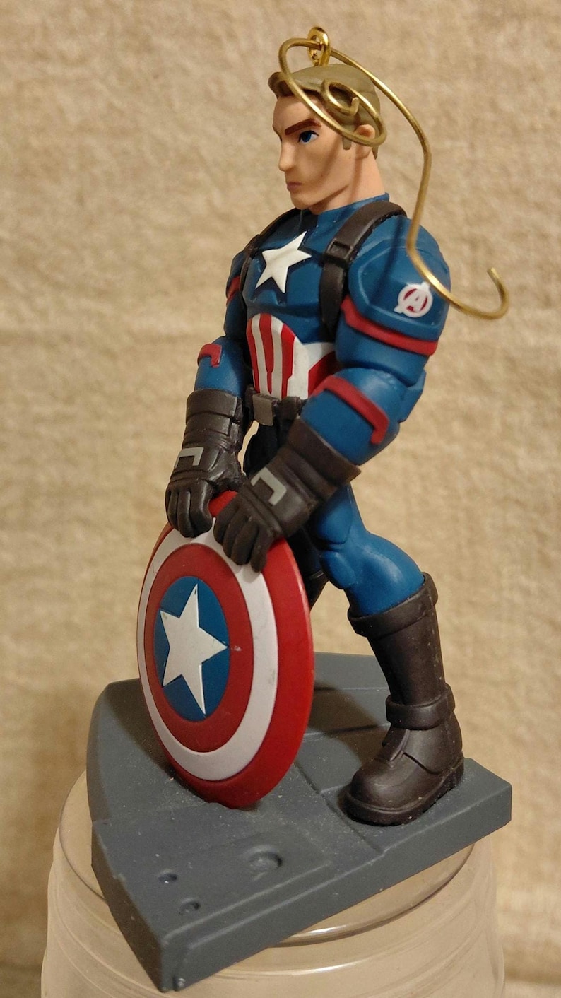 Puede incluir: Figura de Capit&aacute;n Am&eacute;rica, con un traje azul y rojo y una estrella blanca. La figura sostiene un escudo con una estrella blanca sobre un c&iacute;rculo azul, rodeado de anillos rojos y blancos. Con un lazo dorado para colgar.
