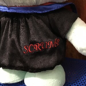 May include: A light blue plush toy wearing a black graduation gown with a blue sash and a red tassel. The gown has the word "SCRUMP" embroidered on the front in red thread.