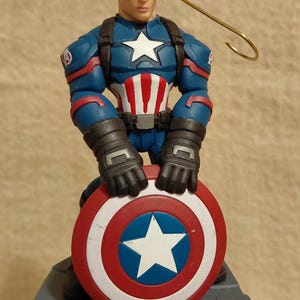 Puede incluir: Figura de Capit&aacute;n Am&eacute;rica sosteniendo su escudo ic&oacute;nico. La figura viste un traje azul, rojo y blanco, con una estrella en el pecho y el escudo. En la parte superior hay un lazo de alambre dorado.