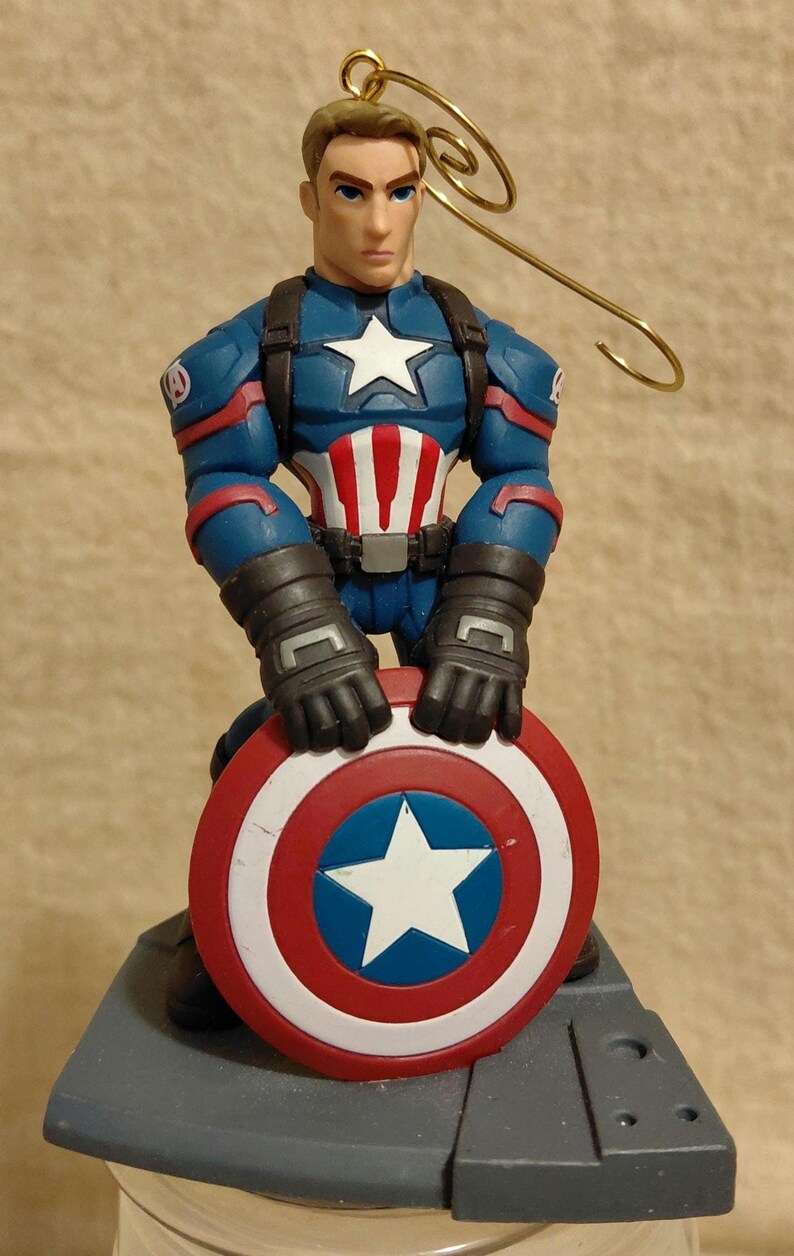 Puede incluir: Figura de Capit&aacute;n Am&eacute;rica sosteniendo su escudo ic&oacute;nico. La figura viste un traje azul, rojo y blanco, con una estrella en el pecho y el escudo. En la parte superior hay un lazo de alambre dorado.