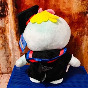 May include: A plush toy wearing a black graduation gown and cap. The toy has a light blue body, yellow hair, and a red and blue sash.