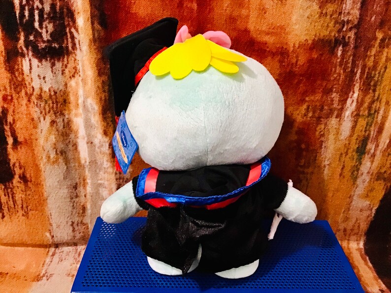 May include: A plush toy wearing a black graduation gown and cap. The toy has a light blue body, yellow hair, and a red and blue sash.