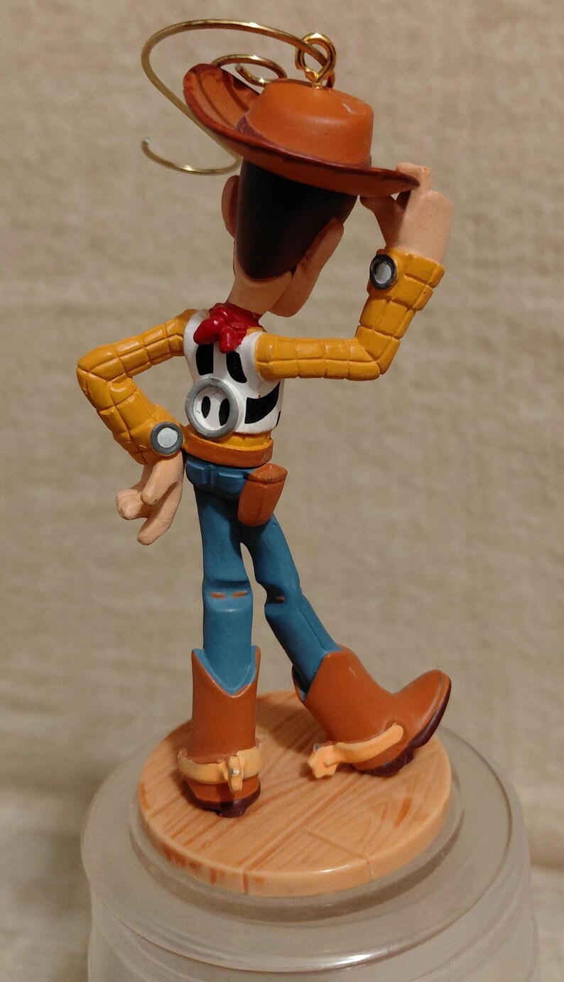 May include: A figurine of the character Woody from the animated film Toy Story. Woody is wearing a brown cowboy hat, yellow shirt, blue jeans, and brown boots. The figurine is standing on a wooden-look base.