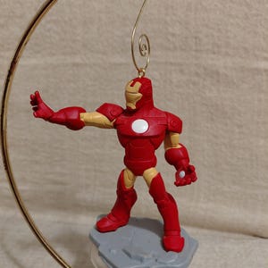May include: A red and gold Iron Man figurine with a white circle on the chest, mounted on a gray base. The figurine is suspended from a gold-colored metal hanger. The figure is in a dynamic pose, with one arm outstretched.