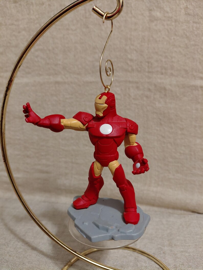 May include: A red and gold Iron Man figurine with a white circle on the chest, mounted on a gray base. The figurine is suspended from a gold-colored metal hanger. The figure is in a dynamic pose, with one arm outstretched.
