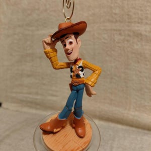 May include: A Woody figurine from the Toy Story franchise. The figure is wearing a brown cowboy hat, yellow shirt, blue pants, and brown boots. The figurine is standing on a wooden-look base with a clear plastic ring and a gold-colored wire for hanging.