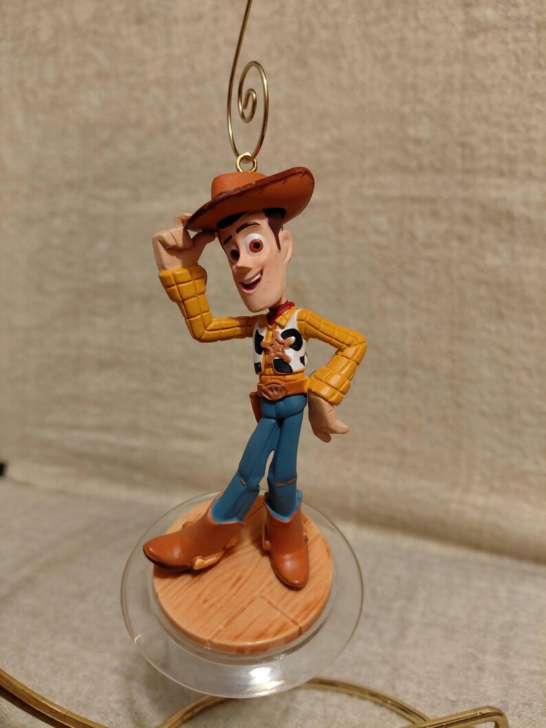 May include: A Woody figurine from the Toy Story franchise. The figure is wearing a brown cowboy hat, yellow shirt, blue pants, and brown boots. The figurine is standing on a wooden-look base with a clear plastic ring and a gold-colored wire for hanging.
