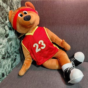 Könnte beinhalten: Ein brauner Plüschbär mit einem roten Basketballtrikot mit der Nummer 23 in Weiß, einem roten Stirnband und schwarz-weißen Turnschuhen. Der Bär liegt auf einer grauen Oberfläche.