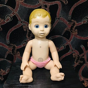 May include: A vintage baby doll with blonde hair, blue eyes, and a pink diaper. The doll is sitting with its legs spread. The background is a black fabric with a repeating pattern of brown belts and horses.