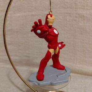 May include: A red Iron Man figurine in a dynamic pose, mounted on a gray base. The superhero figure is suspended from a gold-colored metal hanger. The figurine has white accents and is a collectible item.