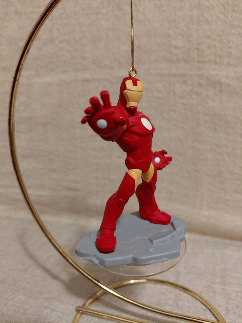 May include: A red Iron Man figurine in a dynamic pose, mounted on a gray base. The superhero figure is suspended from a gold-colored metal hanger. The figurine has white accents and is a collectible item.