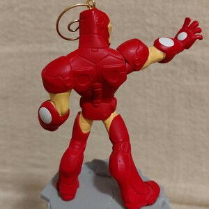 May include: A red Iron Man figurine with a gold loop for hanging. The figure is posed with one arm outstretched, and the other bent. The figure is standing on a gray base. The figure has yellow accents.