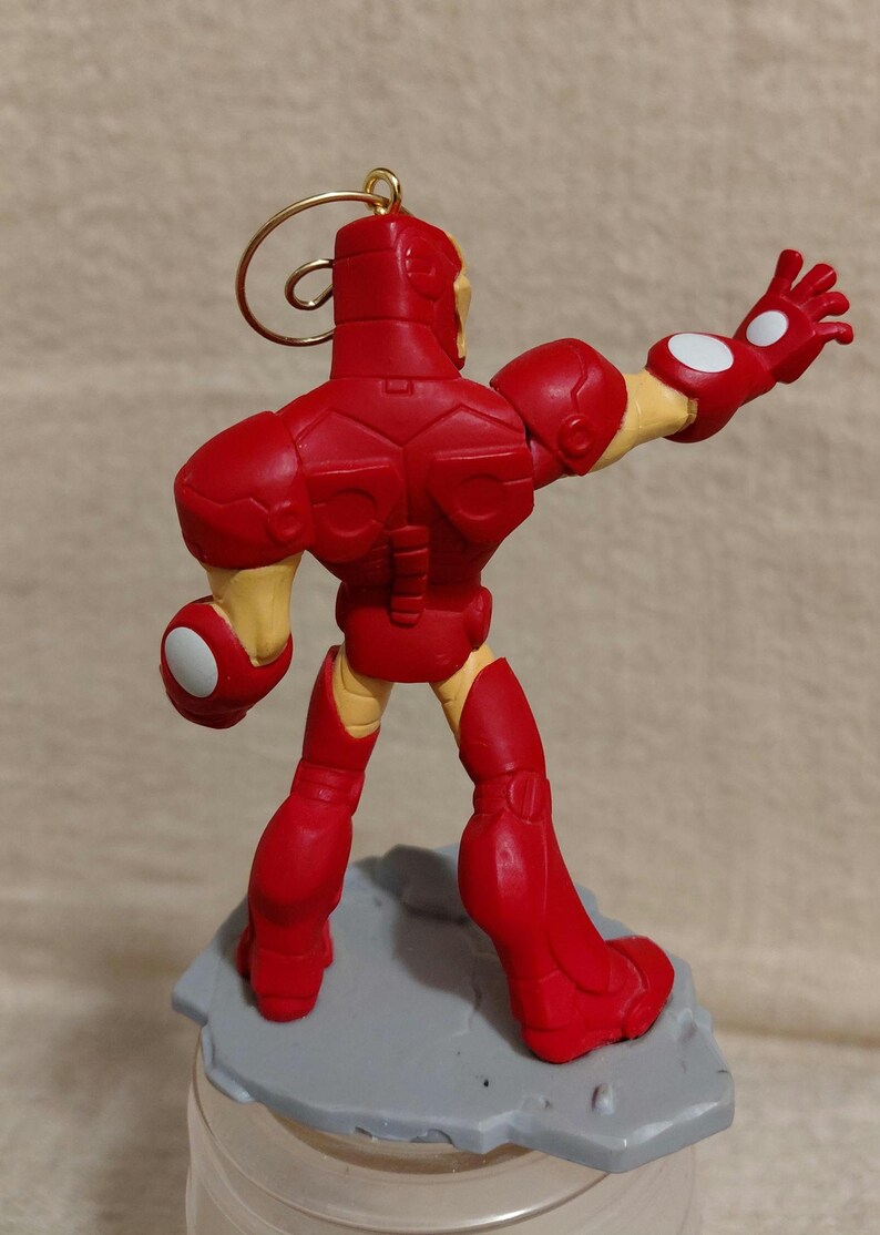 May include: A red Iron Man figurine with a gold loop for hanging. The figure is posed with one arm outstretched, and the other bent. The figure is standing on a gray base. The figure has yellow accents.