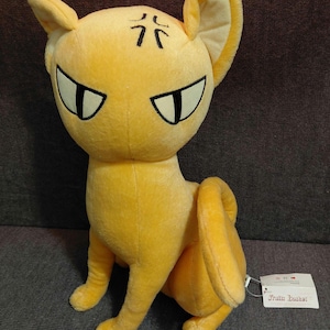 May include: A plush, yellow cat-like character with large, angled eyes and black markings above the eyes. The stuffed animal has large ears and is in a seated position. A tag is attached to the toy.