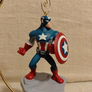 May include: A Captain America ornament figurine. The figurine is in a heroic pose, holding a shield. The figure is wearing a blue and white costume with red boots and gloves. The shield is red, white, and blue with a white star.