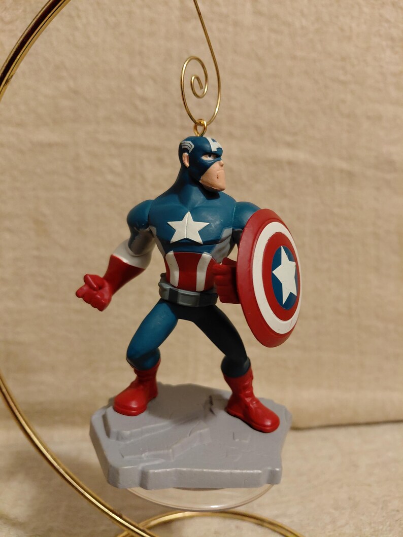 May include: A Captain America ornament figurine. The figurine is in a heroic pose, holding a shield. The figure is wearing a blue and white costume with red boots and gloves. The shield is red, white, and blue with a white star.