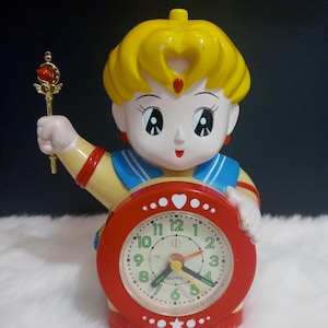 Puede incluir: Un reloj despertador rojo y amarillo con forma de personaje de dibujos animados con cabello rubio, un cuello marinero azul y una varita dorada. La esfera del reloj tiene un fondo blanco con números y manecillas negros. La esfera del reloj también tiene un corazón rojo y una estrella.