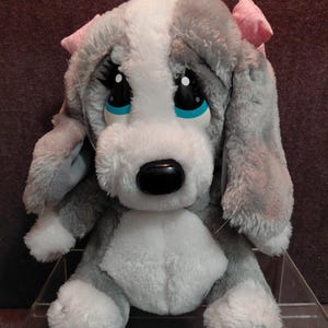 Puede incluir: Un peluche de cachorro con una expresión triste. El perro tiene pelaje gris y blanco, ojos negros grandes con detalles azules y un lazo rosa en la oreja. El juguete está diseñado para parecerse a un perro de dibujos animados.