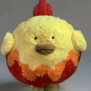 Puede incluir: Un peluche de pollo con un cuerpo amarillo redondo, pequeñas alas y una cresta roja. La parte inferior del pollo es roja con detalles en naranja y amarillo. Tiene un pico marrón y ojos negros pequeños.