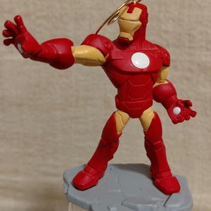 May include: A red and gold Iron Man figurine in a dynamic pose, with one arm extended. The figurine has a white circle on its chest and is mounted on a gray base. A gold wire loop is attached to the head.
