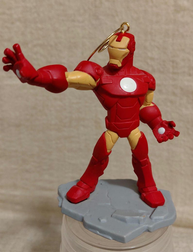 May include: A red and gold Iron Man figurine in a dynamic pose, with one arm extended. The figurine has a white circle on its chest and is mounted on a gray base. A gold wire loop is attached to the head.