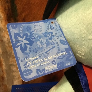 May include: Blue tag with white flowers and a surfboard design. The tag says "Licensee: CV CIPTA ALAM MANDIRI. MADE IN CHINA. &copy;Disney"