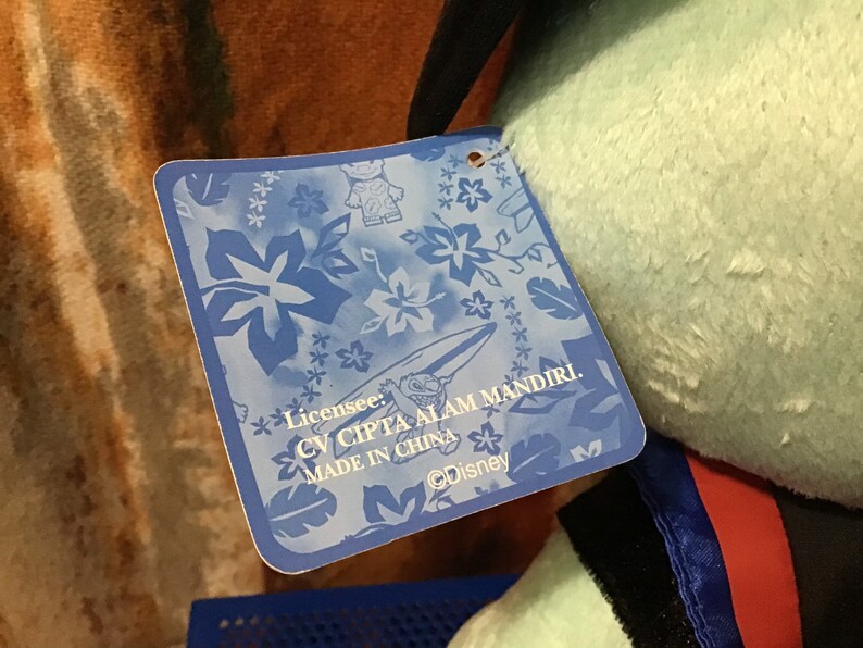 May include: Blue tag with white flowers and a surfboard design. The tag says "Licensee: CV CIPTA ALAM MANDIRI. MADE IN CHINA. &copy;Disney"