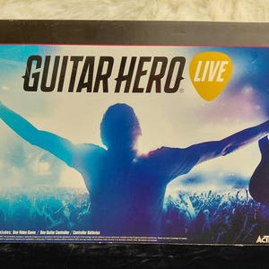 Pode incluir: Caixa do jogo Guitar Hero Live para PS3. A caixa apresenta uma silhueta de uma pessoa com os braços estendidos em frente a uma multidão. O título do jogo está em letras grandes com um gráfico de palheta amarelo.