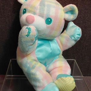 May include: A pastel-colored plush bear with a white face, pink nose, and teal eyes. The bear has teal satin accents on its paws and chest. The left paw has the embroidered text "Missy's Toy". The bear is sitting on a clear acrylic surface.