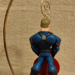 Puede incluir: Una figura de superh&eacute;roe con un traje azul y rojo, de pie sobre una base gris. La figura est&aacute; unida a un colgador de adorno de metal dorado. El superh&eacute;roe tiene una capa roja y botas negras.
