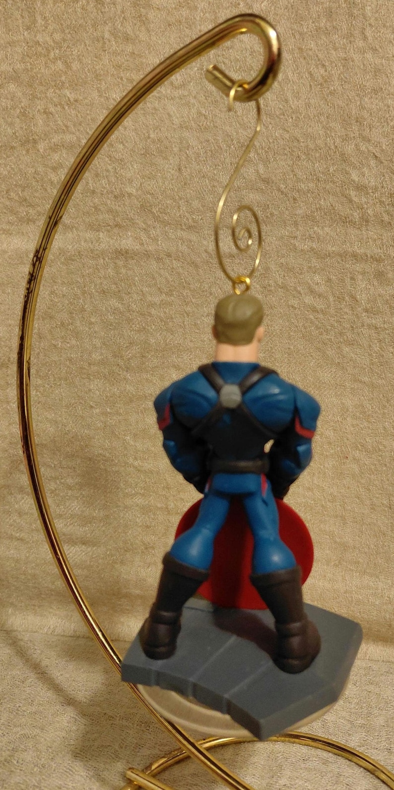 Puede incluir: Una figura de superh&eacute;roe con un traje azul y rojo, de pie sobre una base gris. La figura est&aacute; unida a un colgador de adorno de metal dorado. El superh&eacute;roe tiene una capa roja y botas negras.