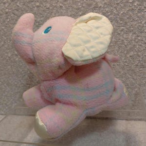 May include: A plush pink elephant toy with pastel stripes and a cream-colored quilted ear. The elephant has a blue eye and a white satin foot. A soft toy for children.