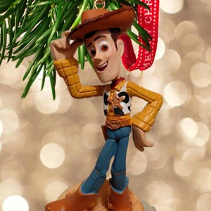 May include: A Woody Christmas ornament. The figure is wearing a brown cowboy hat, yellow shirt, blue jeans, and brown boots. The ornament is hanging from a Christmas tree branch with a red ribbon that says "Merry Christmas".