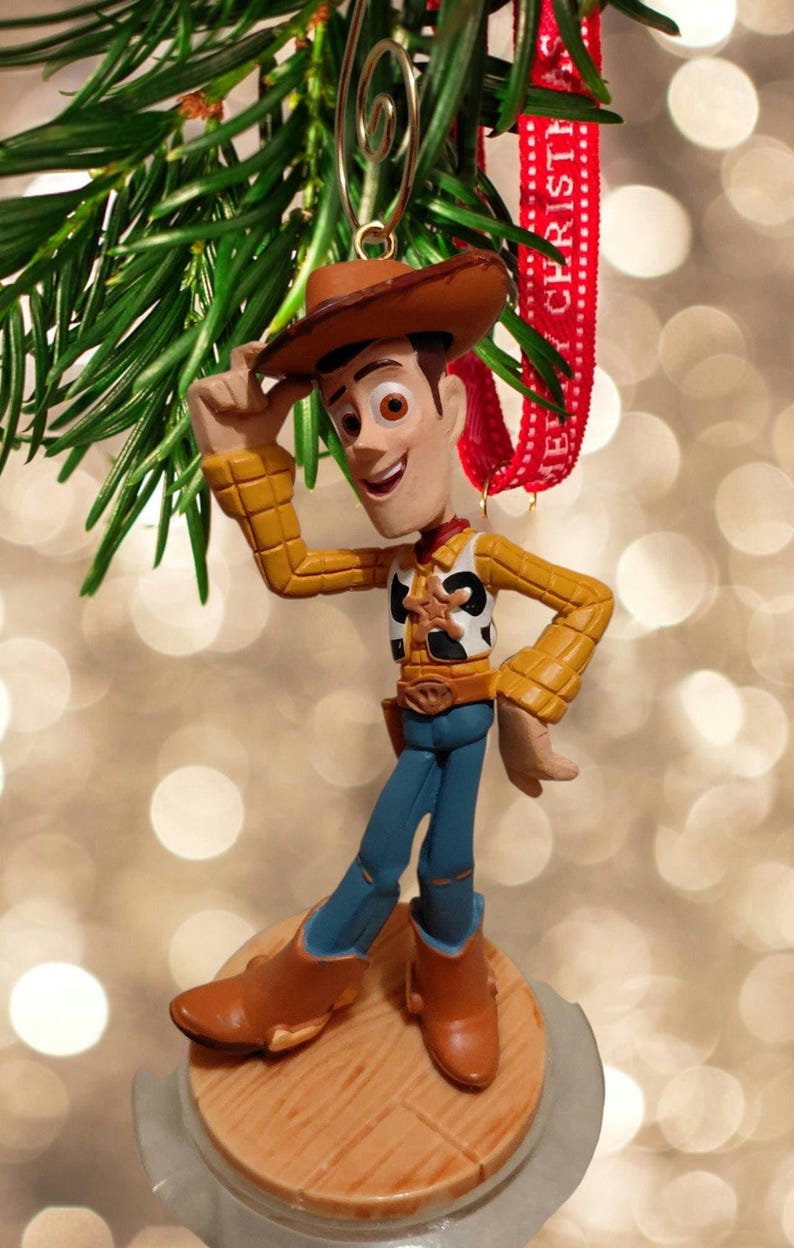 May include: A Woody Christmas ornament. The figure is wearing a brown cowboy hat, yellow shirt, blue jeans, and brown boots. The ornament is hanging from a Christmas tree branch with a red ribbon that says "Merry Christmas".