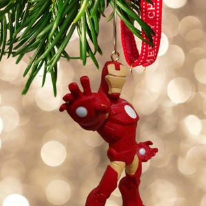 May include: A red and gold Iron Man Christmas ornament. The superhero figure is posed with one arm outstretched. The ornament hangs from a green pine branch and a red ribbon with the text "MERRY CHRISTMAS".