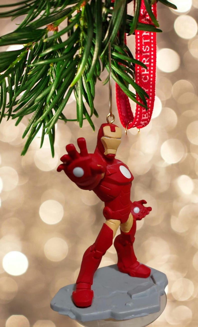 May include: A red and gold Iron Man Christmas ornament. The superhero figure is posed with one arm outstretched. The ornament hangs from a green pine branch and a red ribbon with the text "MERRY CHRISTMAS".