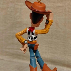 May include: A Woody figurine from the Toy Story franchise. The figure wears a brown cowboy hat, yellow shirt, blue jeans, and brown boots. The ornament has a gold-colored loop for hanging.