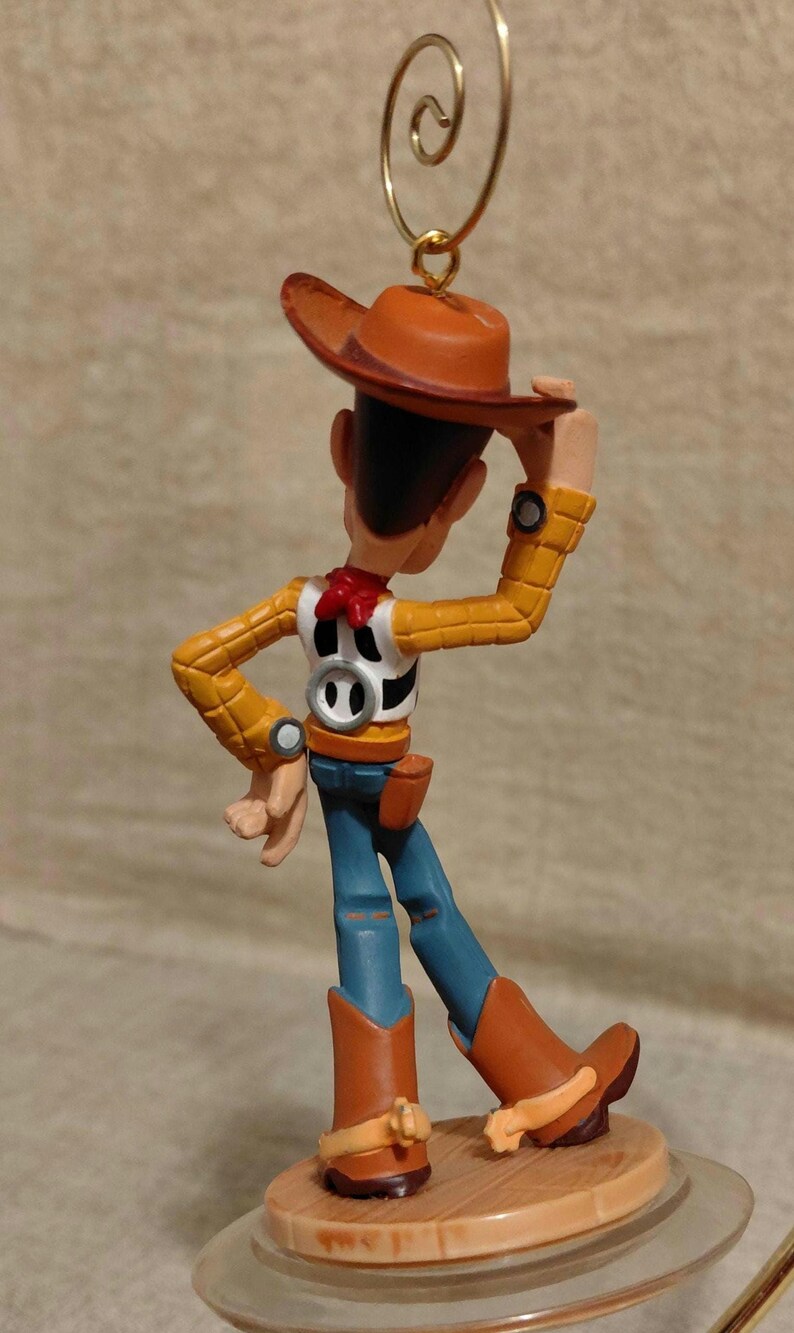 May include: A Woody figurine from the Toy Story franchise. The figure wears a brown cowboy hat, yellow shirt, blue jeans, and brown boots. The ornament has a gold-colored loop for hanging.