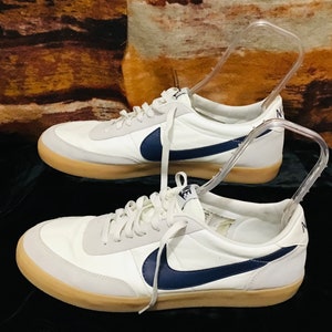 Puede incluir: Un par de zapatillas de lona blancas con un logotipo Swoosh azul marino. Las zapatillas tienen una suela de goma y cordones blancos.