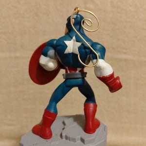 May include: A Captain America figurine, viewed from the back, stands on a gray base. The figure wears a blue suit with a white star, red boots, and gloves. A red shield is held on the left arm. A gold wire loop is attached to the head.