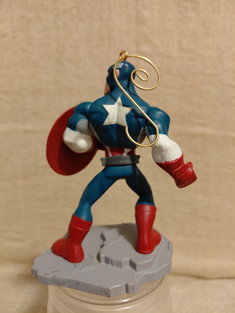 May include: A Captain America figurine, viewed from the back, stands on a gray base. The figure wears a blue suit with a white star, red boots, and gloves. A red shield is held on the left arm. A gold wire loop is attached to the head.