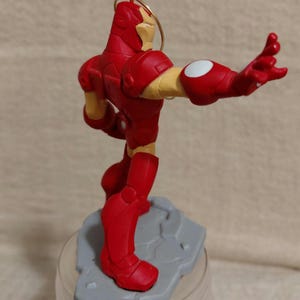 May include: A red and gold Iron Man figurine, posed with one arm extended. The figurine is on a gray base and has a gold loop for hanging. The figure's arm has a white circle on the elbow. The figurine is a collectible item.